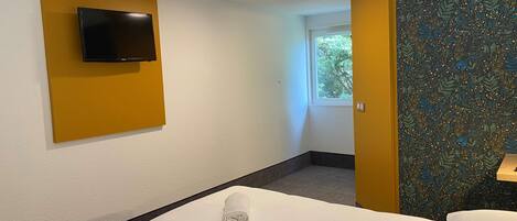 Habitación de hotel con cama, televisión montada en una pared amarilla y ventana con vista a árboles.