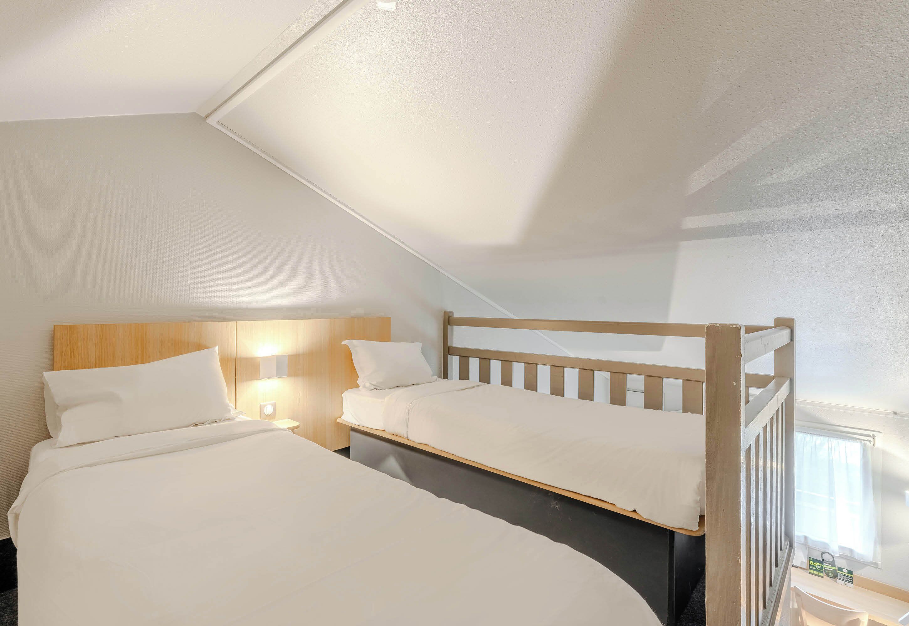 A bedroom with two beds, a wooden headboard, a nightstand with a lamp, and a window with curtains.