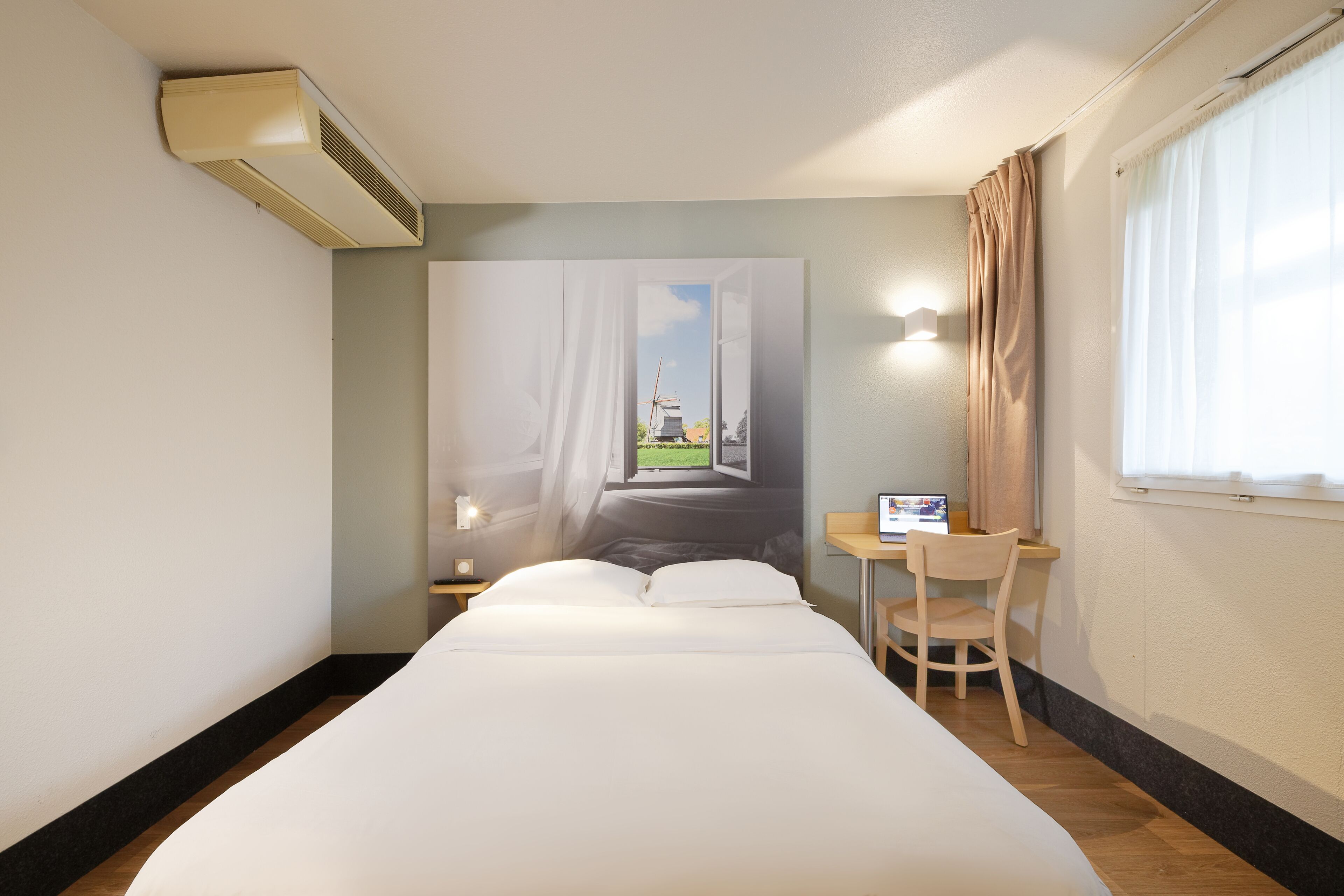 A hotel room with a bed, a desk, a chair, a window with curtains, and an air conditioning unit.