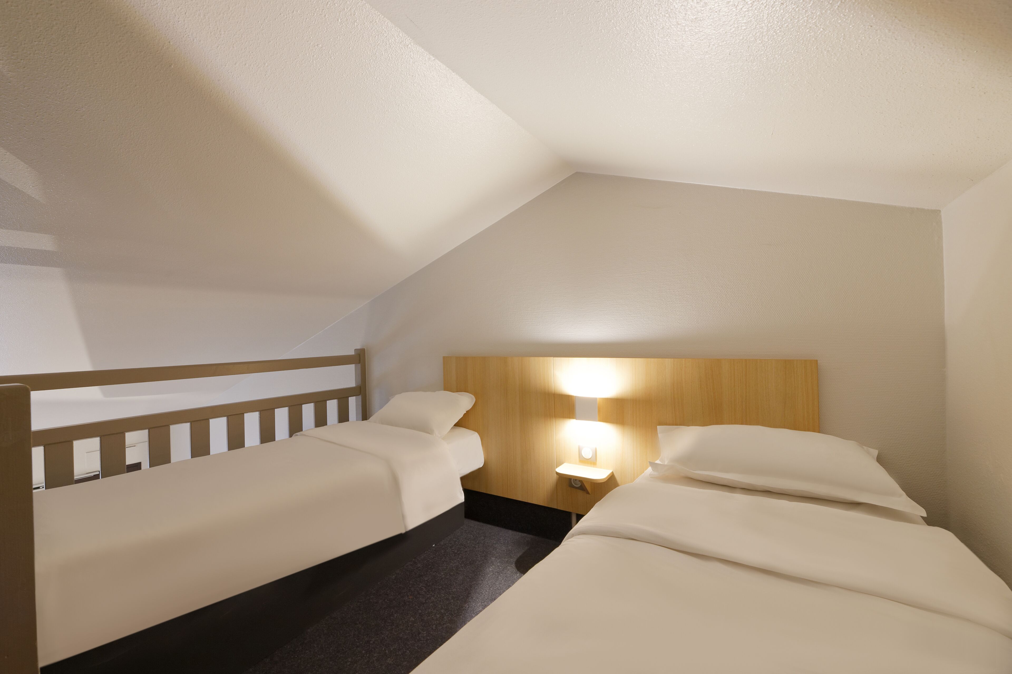 A hotel room with two single beds, a wooden headboard, a nightstand with a lamp, and a carpeted floor.