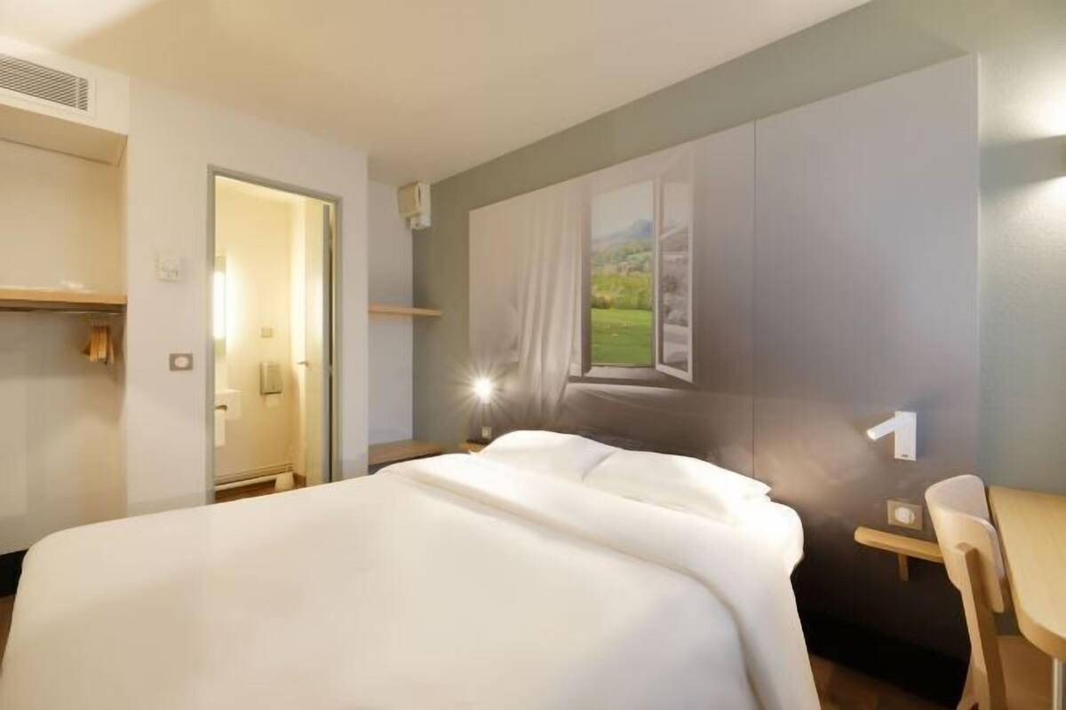 A hotel room with a bed, a desk, a chair, a window with a view, and a door.