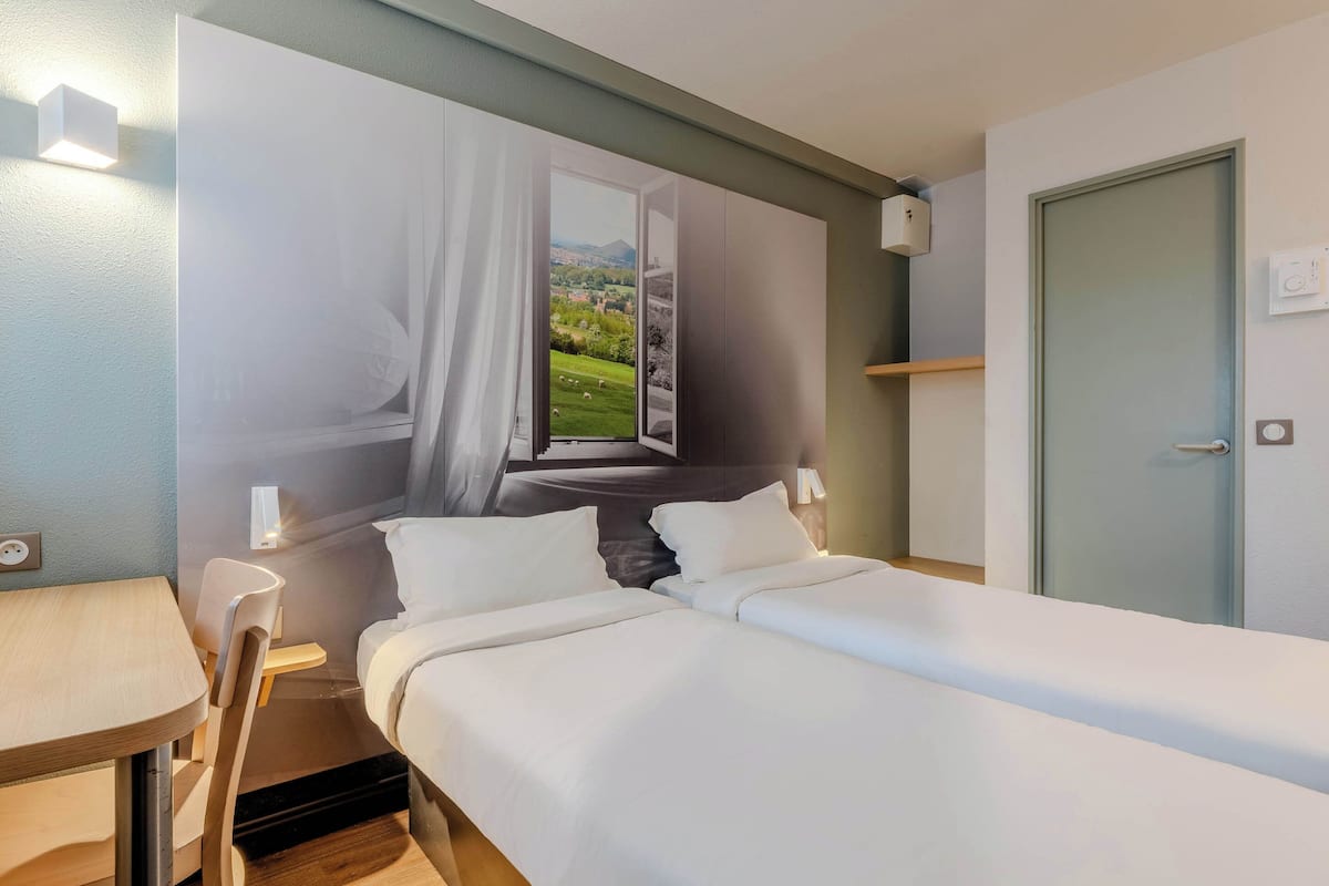 A hotel room with a bed, a desk, and a chair. There is a window with a view of a green landscape.