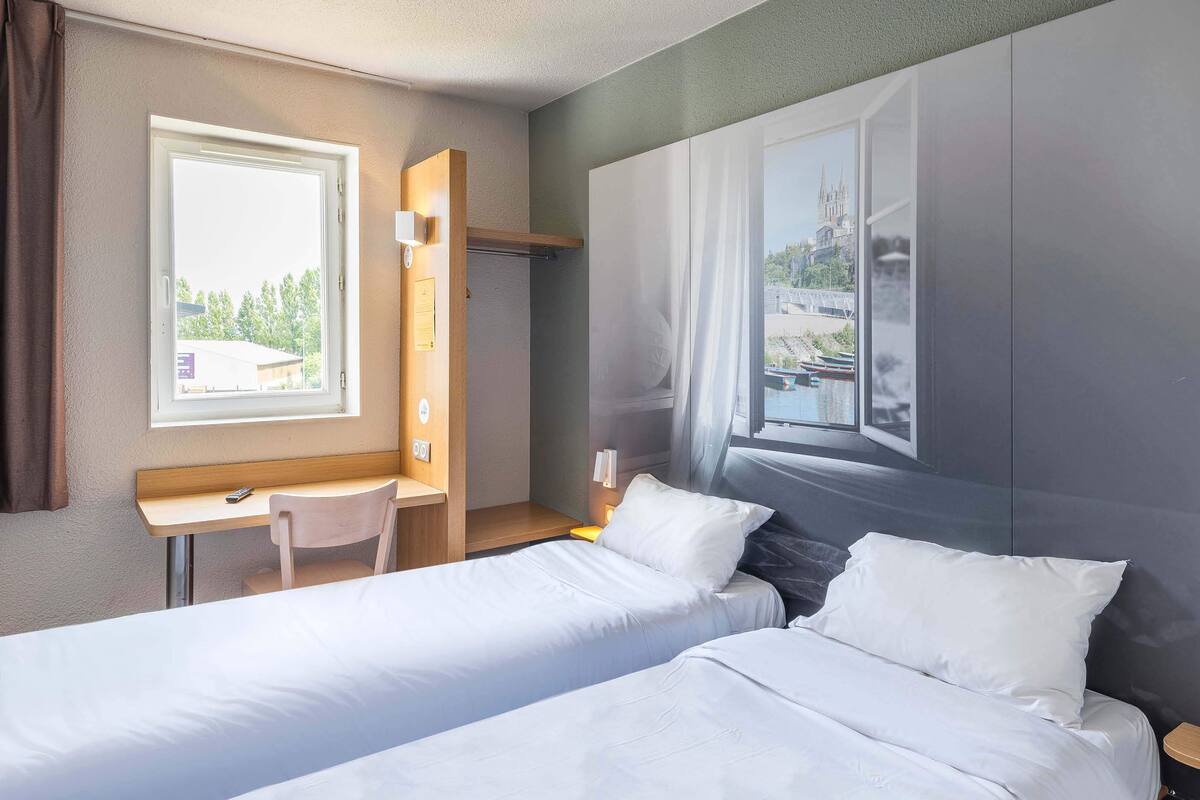 A hotel room with two single beds, a desk, and a chair. There is a window with a view of a building and trees.