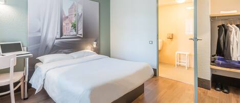 A modern hotel room with a large bed, a desk with a laptop, a wardrobe, and a view of a building through a window.