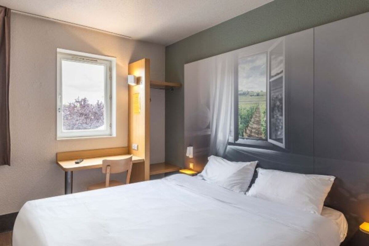 Habitación de hotel con cama, escritorio y silla. Se ve una ventana con vistas a árboles y un edificio.