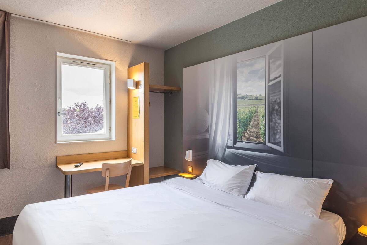 A hotel room with a bed, a desk, a chair, and a large window with a view of a field.