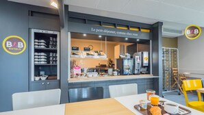 Un área de desayuno con mostrador que exhibe pasteles y una máquina de café, una mesa con artículos para el desayuno, y un cartel que dice "Le petit-déjeuner, comme chez vous".