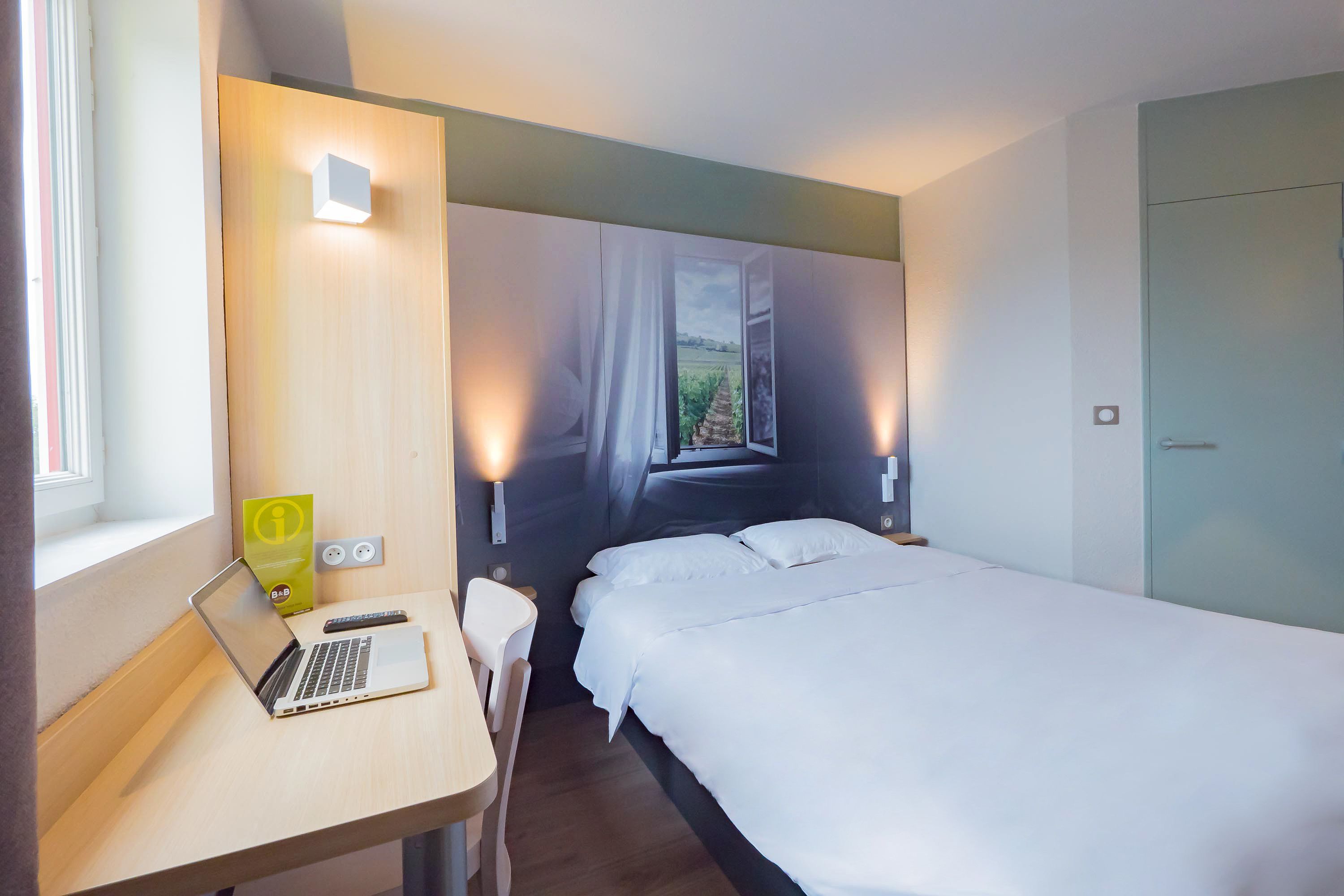 Una habitación de hotel con cama, escritorio con computadora portátil, silla y ventana con cortinas.