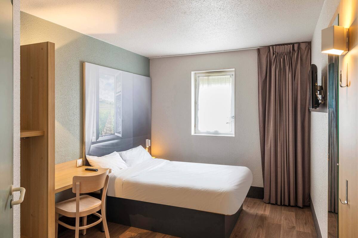 A compact hotel room with a single bed, a desk, a chair, a window, and a curtain.