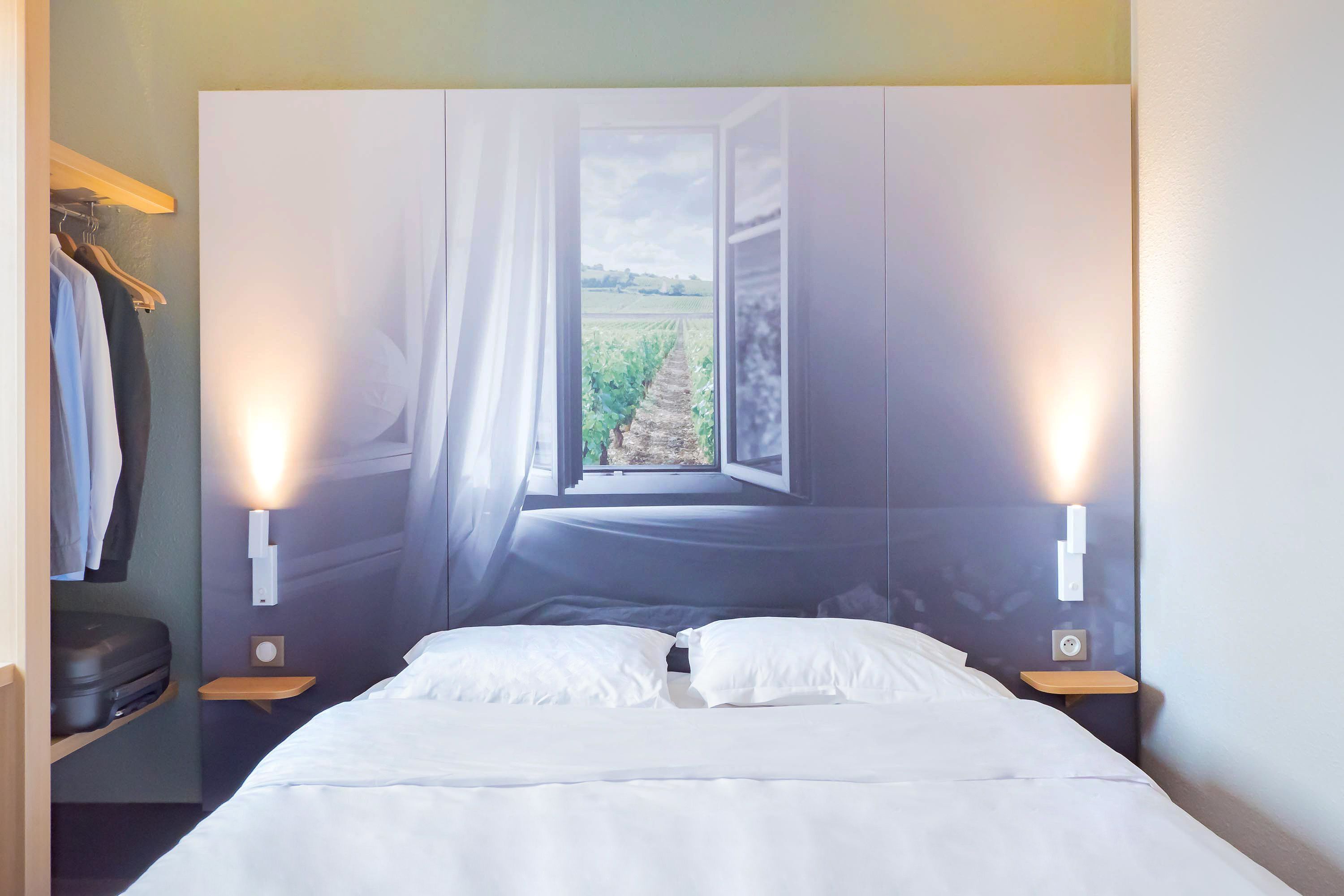 Een slaapkamer met een bed, een nachtkastje, een wandlamp en een raam met uitzicht op een wijngaard.