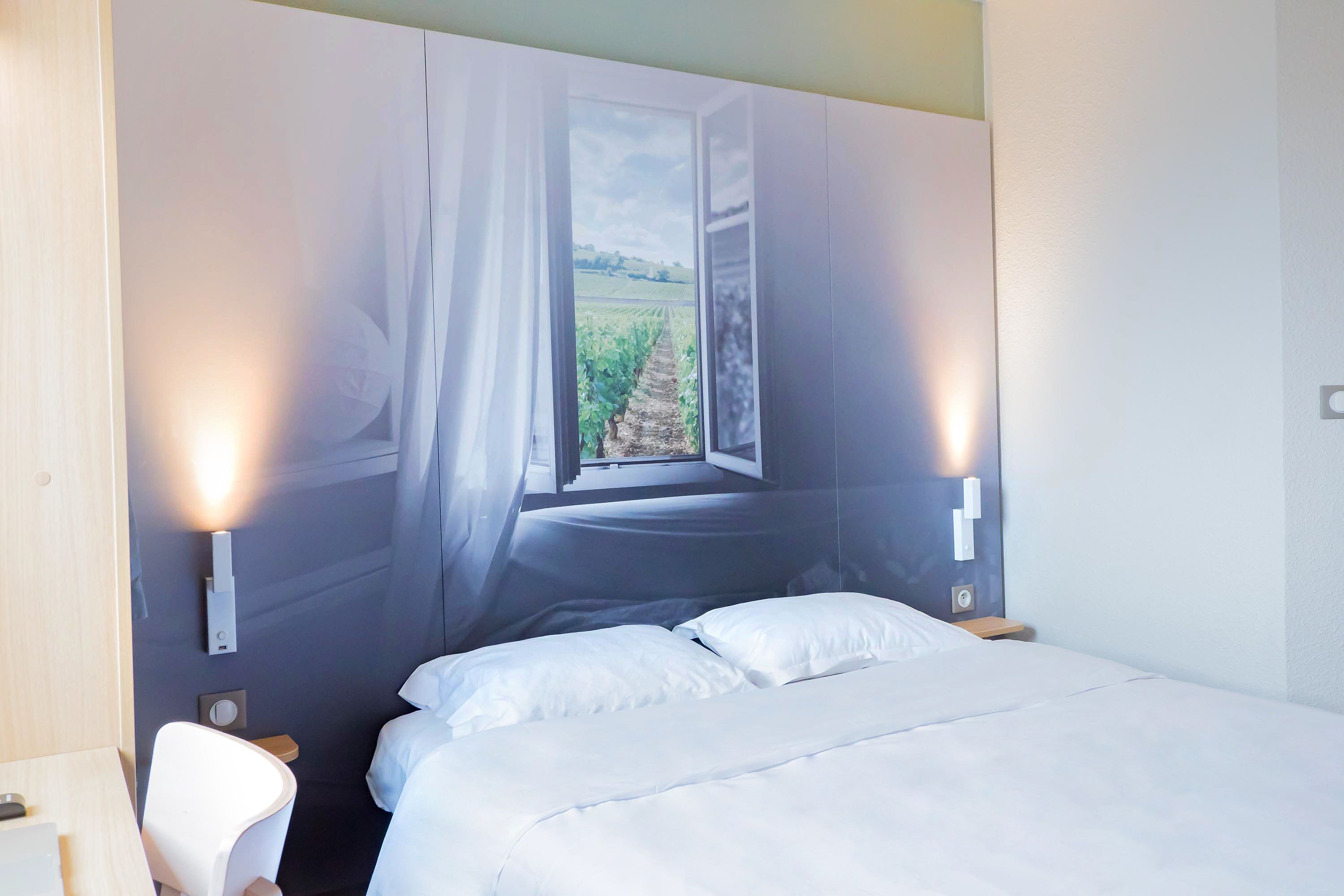 Een slaapkamer met een bed, een stoel, een raam met uitzicht op een wijngaard en wandlampen.