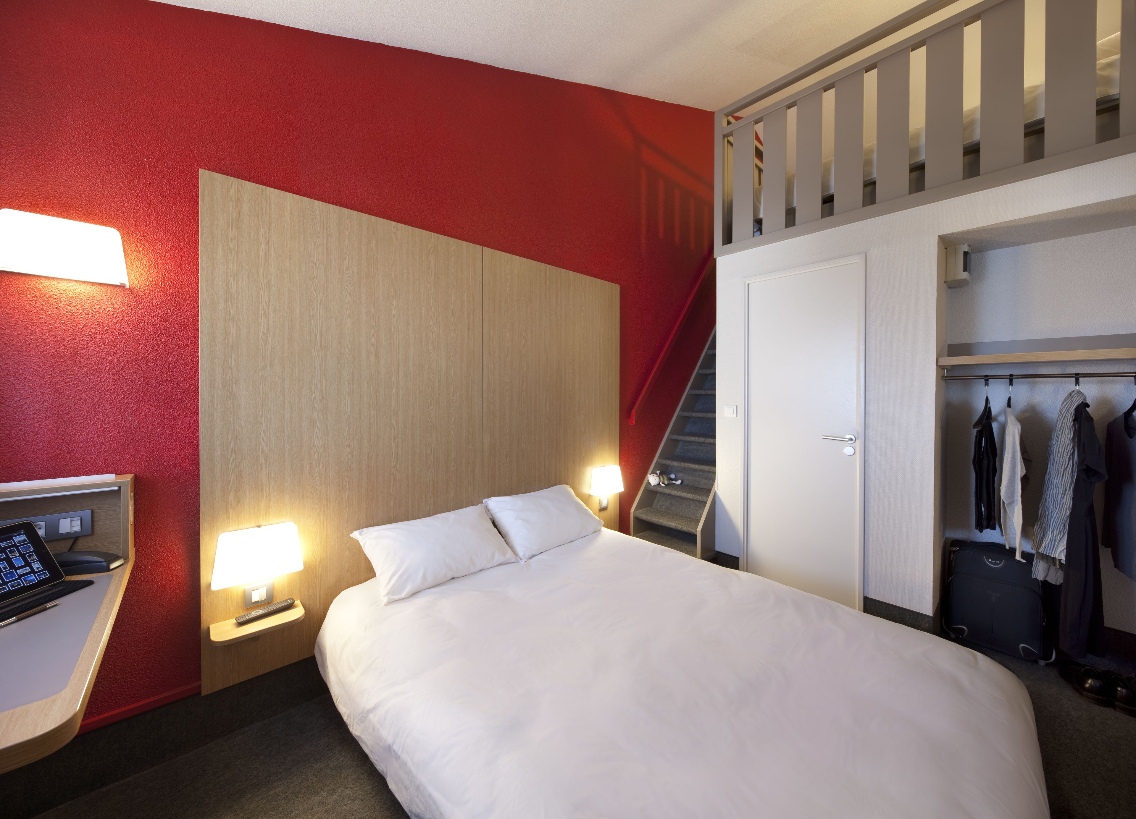 Habitación de hotel con cama, mesitas de noche, escritorio con computadora, lámpara, armario y una escalera.