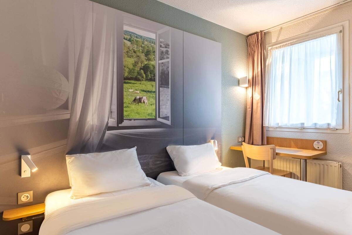 Habitación de hotel con dos camas, un escritorio, una silla, una ventana con cortinas y una lámpara en la pared.