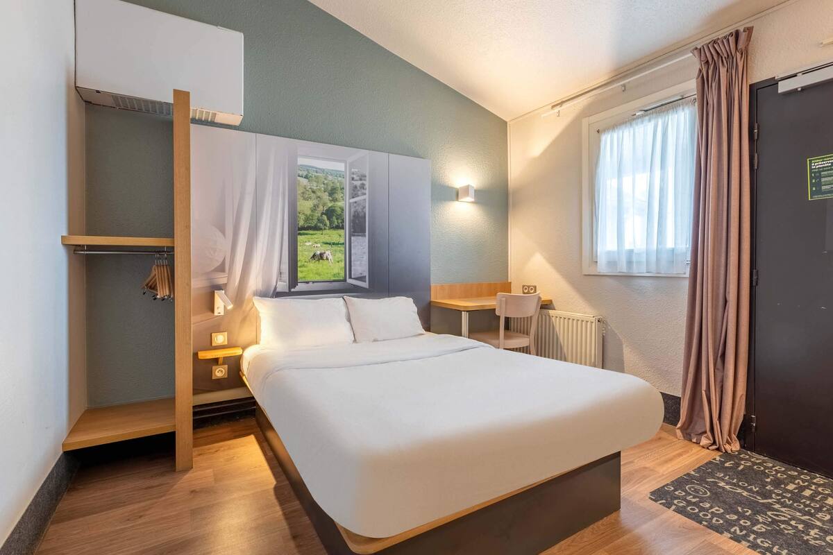 Un dormitorio con cama, escritorio y silla. Se ve una ventana con cortinas, un radiador y una puerta.