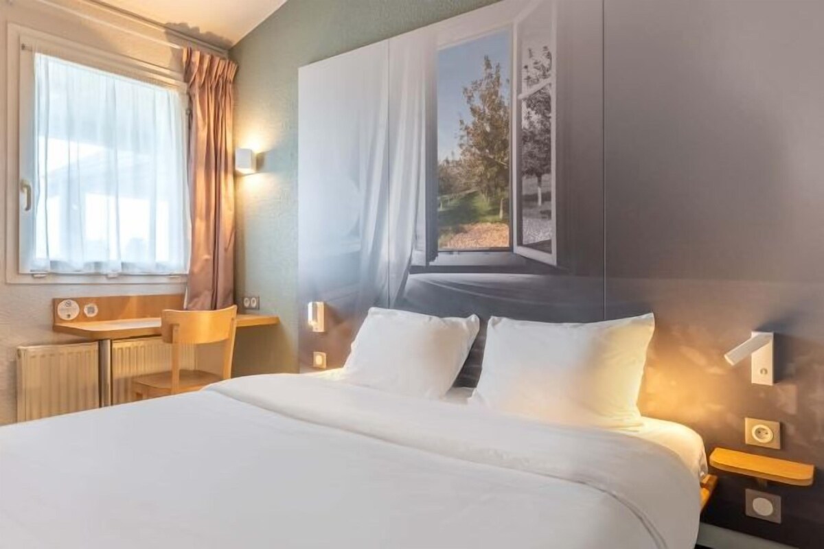 Una habitación de hotel con una cama grande, un escritorio, una silla, una ventana con cortinas y una lámpara de pared.