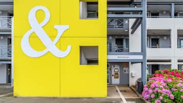 Un edificio amarillo con un gran símbolo de 'y' blanco, un cartel de bienvenida y un cantero con flores rosas.