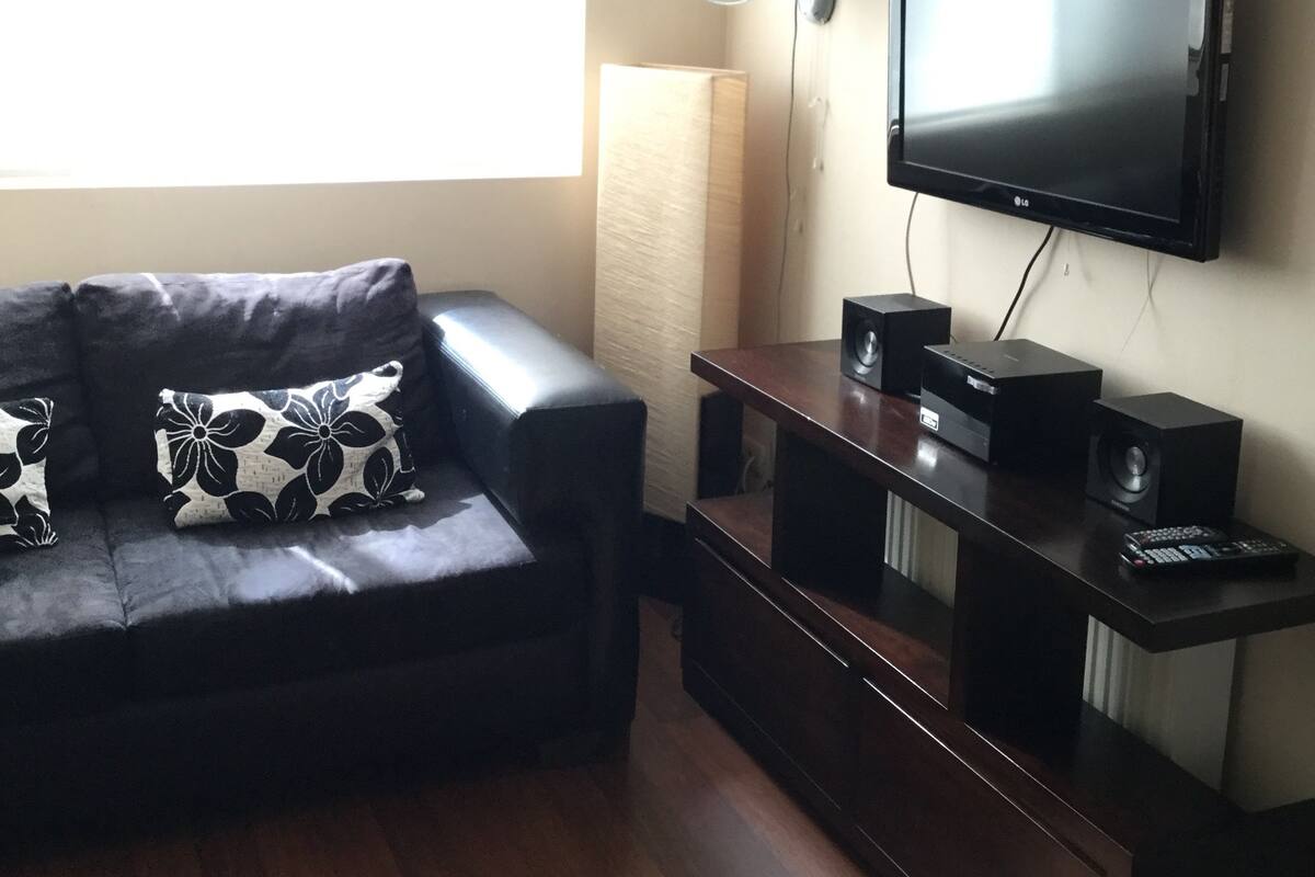 Una sala de estar con un sofá de cuero negro, un televisor de pantalla plana montado en la pared, una lámpara de pie y un mueble de entretenimiento de madera con parlantes.