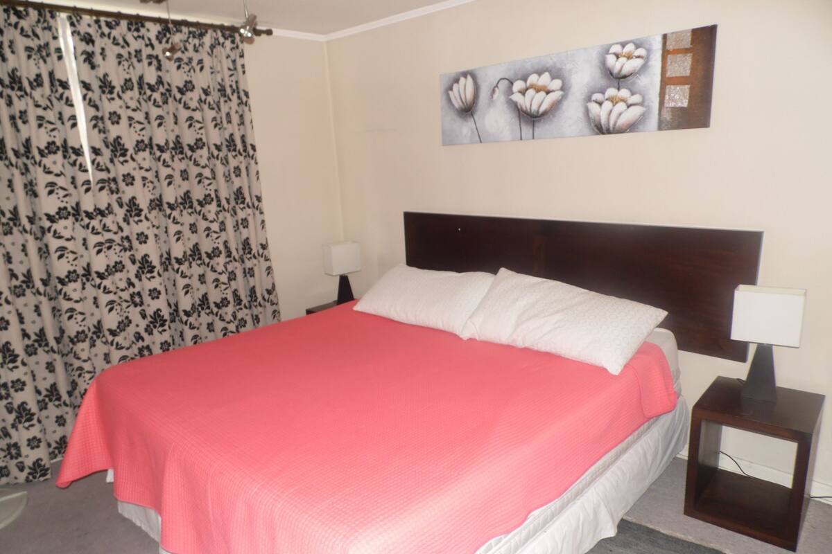 Una cama bien tendida con una colcha rosa, un cabecero de madera, una mesita de noche con una lámpara y una cortina estampada con motivos florales.