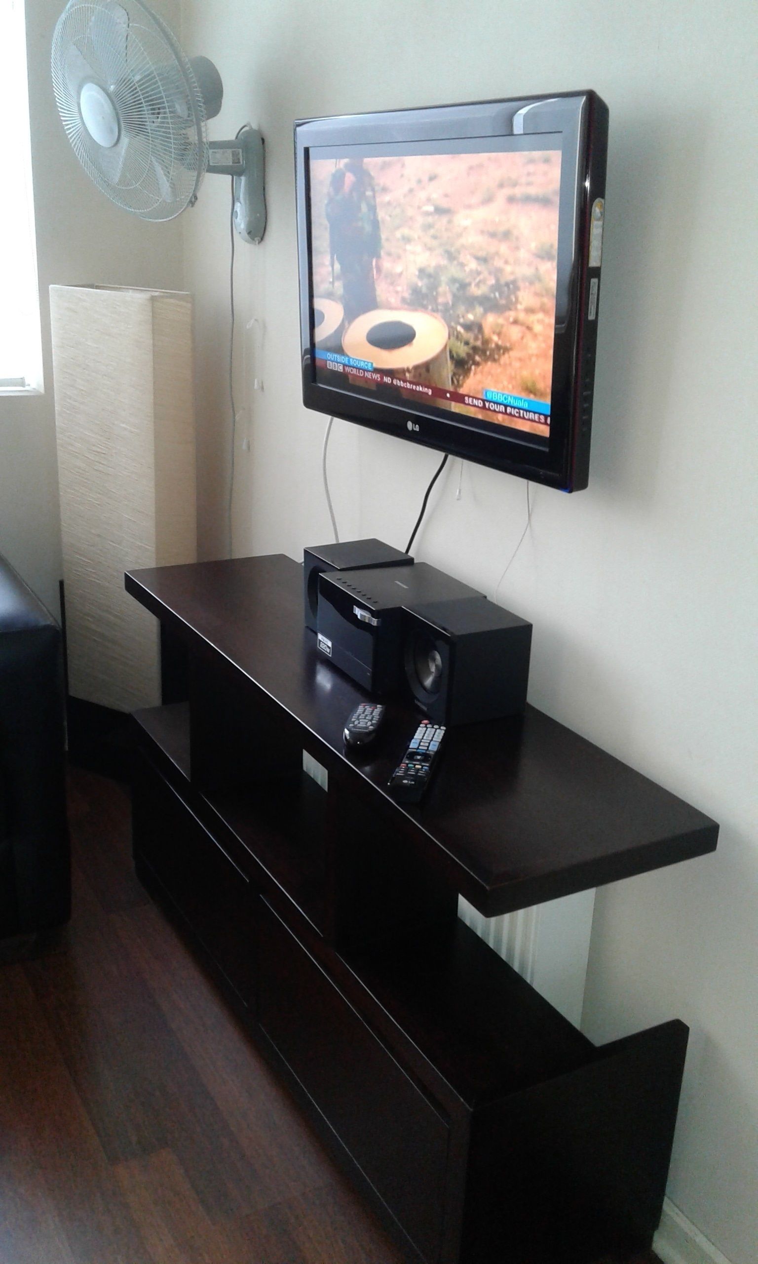 Un televisor de pantalla plana montado en la pared, un mueble de entretenimiento negro debajo y un ventilador de pie a la izquierda.