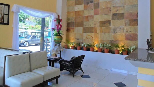 A hotel lobby with a white sofa, a wicker chair, a tiled wall with a checkered pattern, and a row of potted plants. - Hotel Italia (La Ceiba)