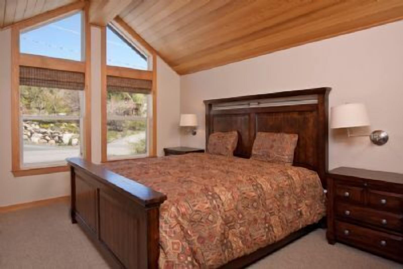 Ein Schlafzimmer mit einem großen Bett, einem hölzernen Kopfteil, zwei Nachttischlampen und Blick ins Freie durch große Fenster.