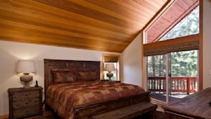Un dormitorio con techo de madera, una cama grande con colcha estampada, dos mesitas de noche con lámparas, un banco al pie de la cama y vistas al exterior a través de una puerta corrediza de cristal.