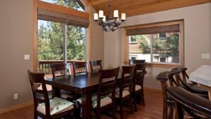 Un comedor con una mesa y sillas de madera, ventanas grandes y una araña.