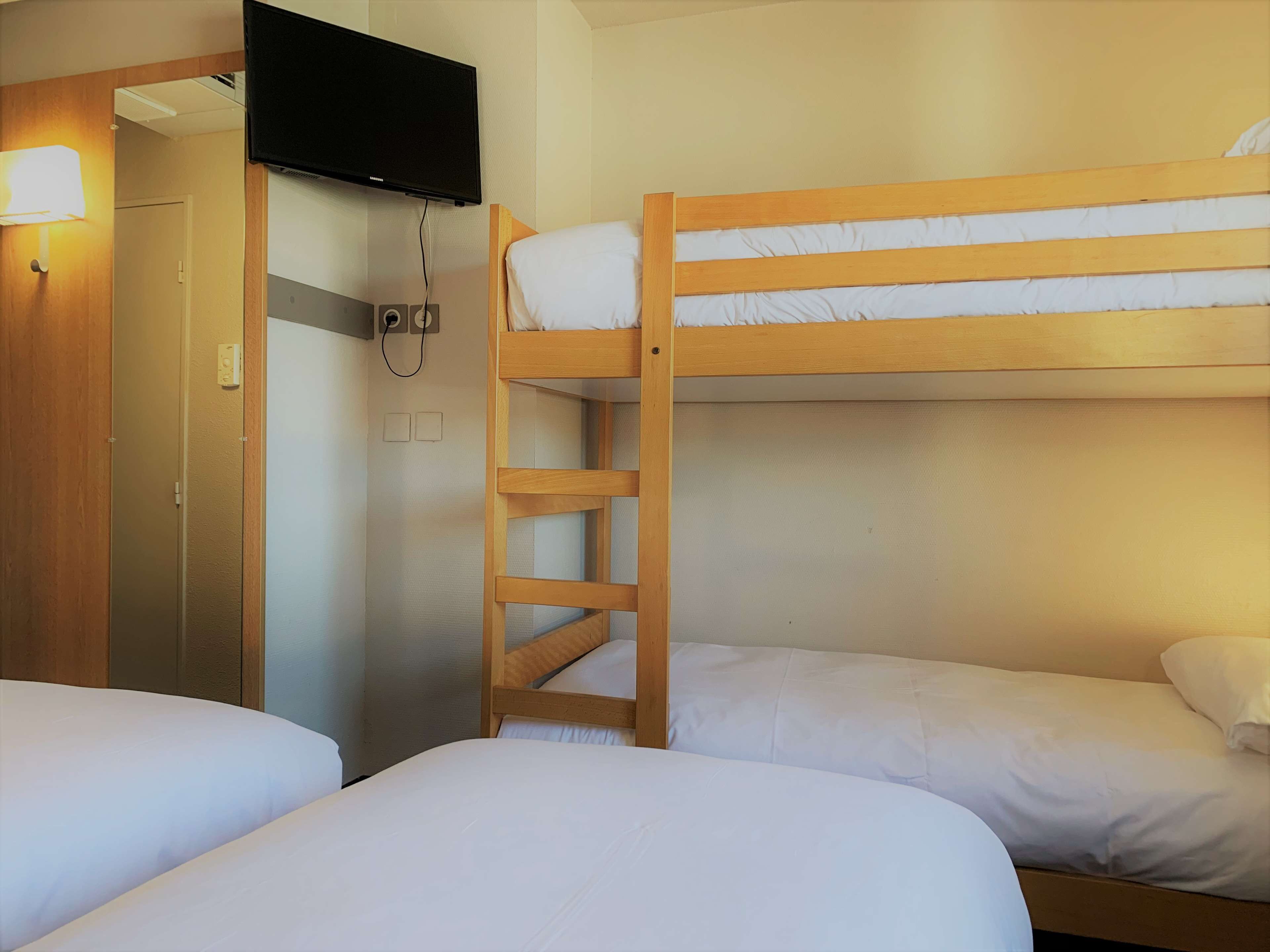 A hotel room with two single beds, a wooden bunk bed, a television mounted on the wall, and a door leading to another room.