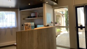 A reception desk with a wooden finish, a window with sheer curtains, and a view of a garden through a glass door. - Kyriad Direct Clermont Ferrand Nord - Gerzat (Gerzat)