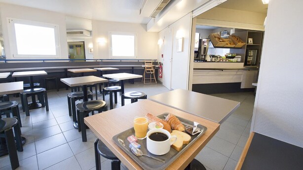 A clean, modern dining area with multiple tables and chairs, a tray with breakfast items, and a kitchen area visible in the background. - Kyriad Direct Clermont Ferrand Nord - Gerzat (Gerzat)