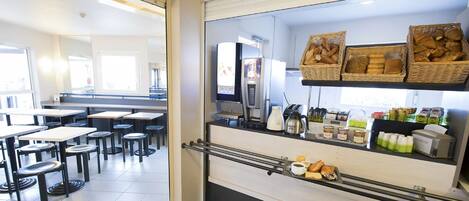 Un café avec des tables et des chaises, une machine à café, ainsi que des paniers remplis de pain et de viennoiseries.