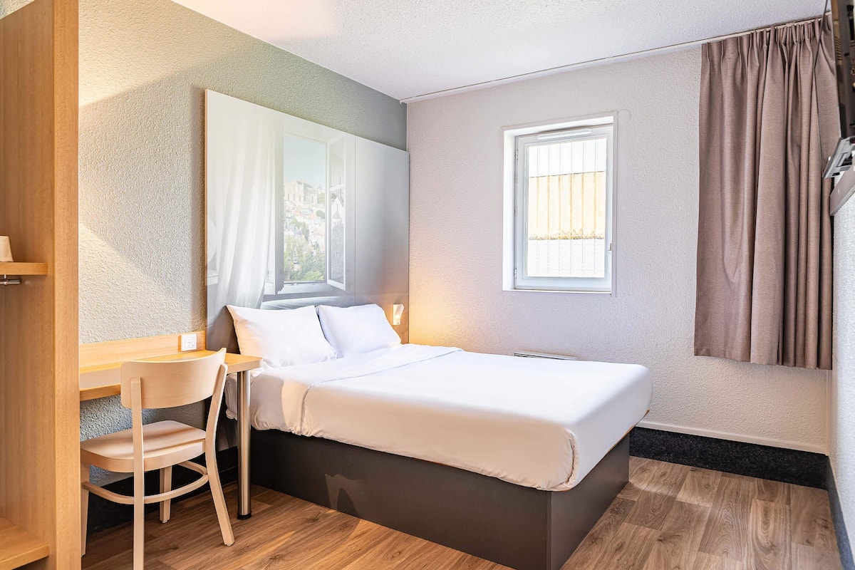 A compact bedroom with a bed, a desk, and a chair, featuring a large mirror and a window with curtains.