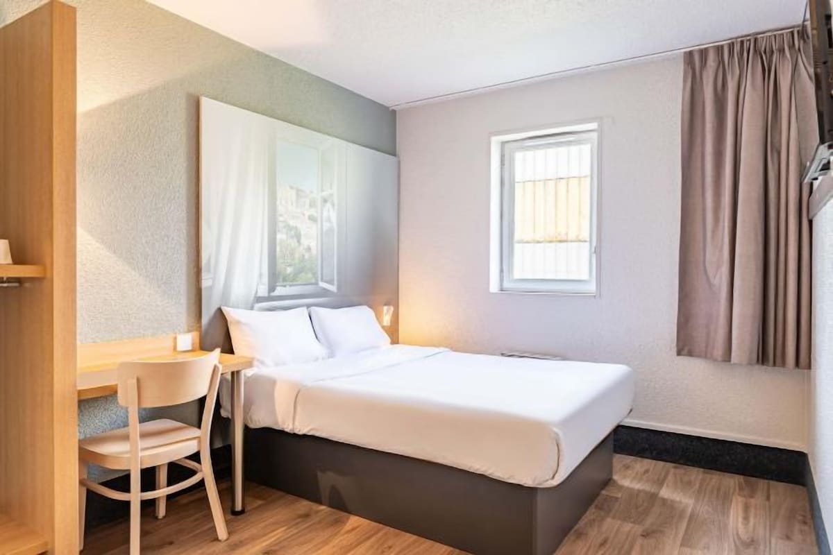 Une chambre d’hôtel de taille réduite, comprenant un lit simple, un petit bureau, une chaise, une fenêtre avec des rideaux et une vue sur l’extérieur.