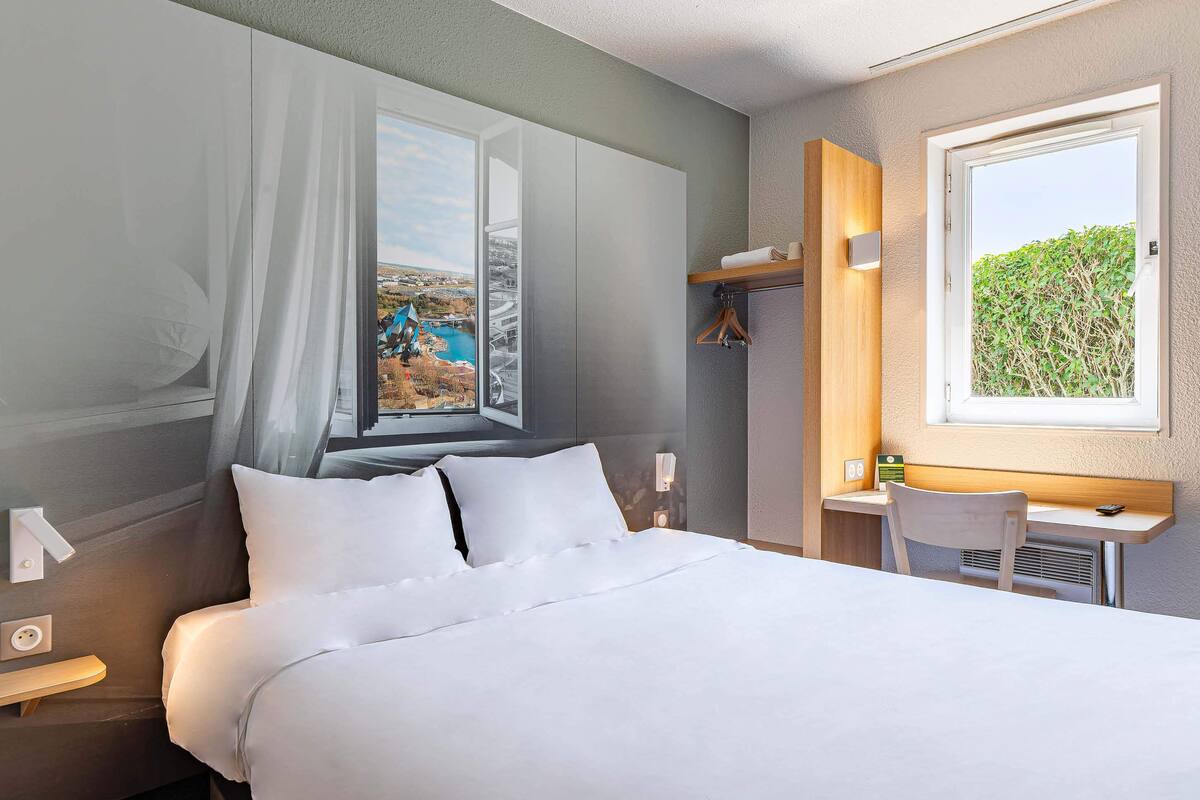 A hotel room with a bed, a desk, a chair, a window with a view of a bridge and greenery, and a hanging coat rack.