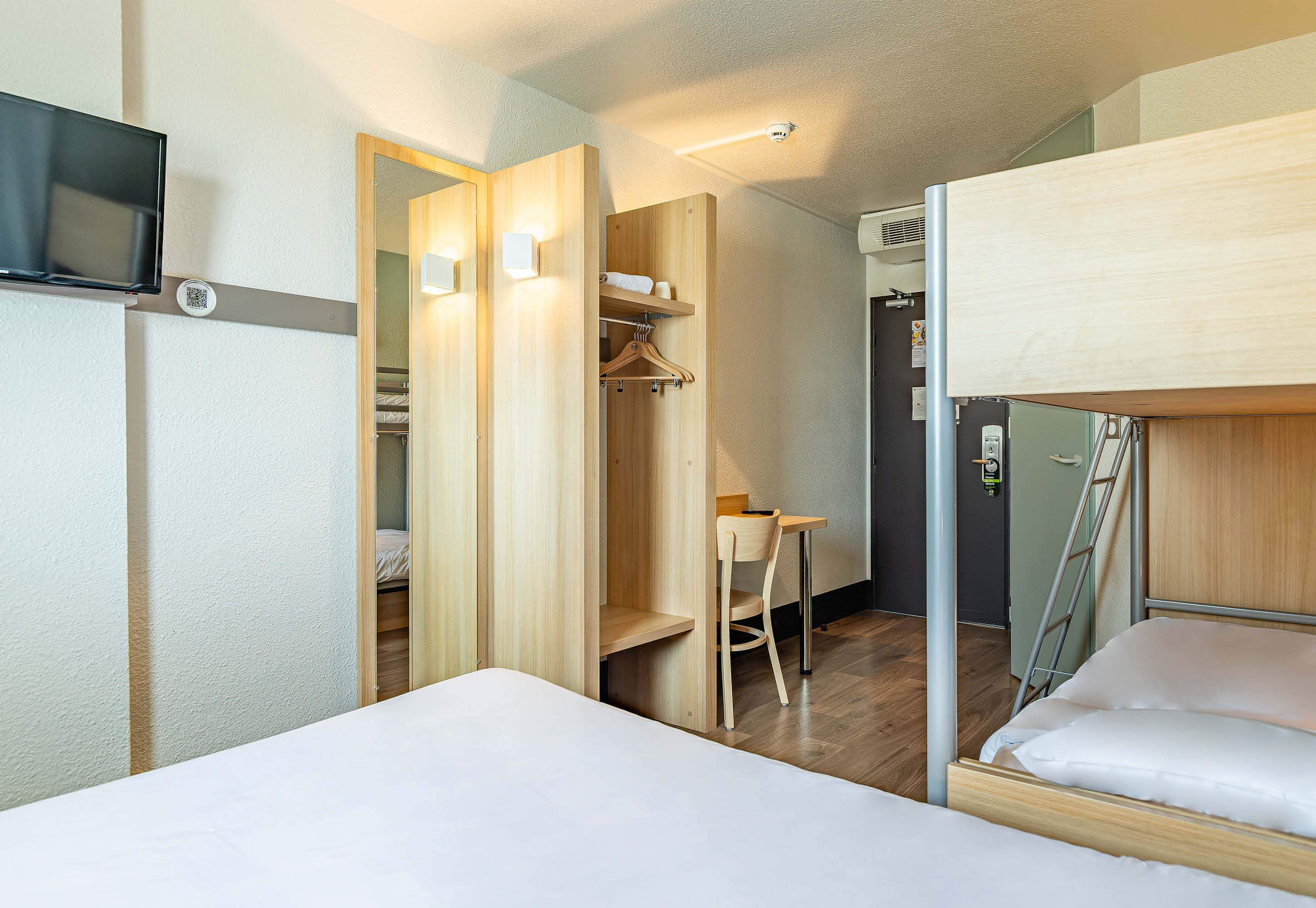 A compact hotel room with a bunk bed, a desk with a chair, a wardrobe, and a television mounted on the wall.