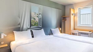 A hotel room with two beds, a window with a view of a cityscape, a desk with a chair, and a small table with a lamp. - B&B HOTEL Poitiers (1) (Chasseneuil-du-Poitou)