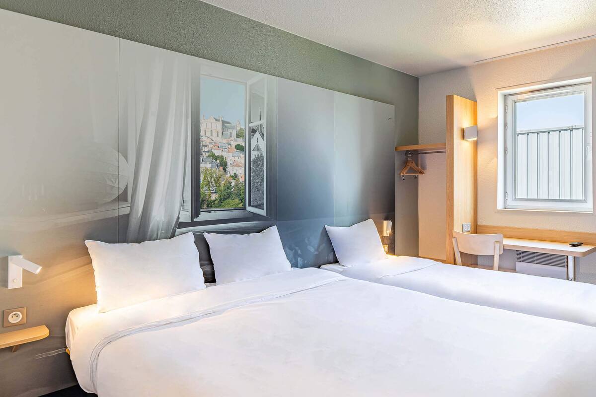 A hotel room with two beds, a window with a view of a cityscape, a desk with a chair, and a small table with a lamp.
