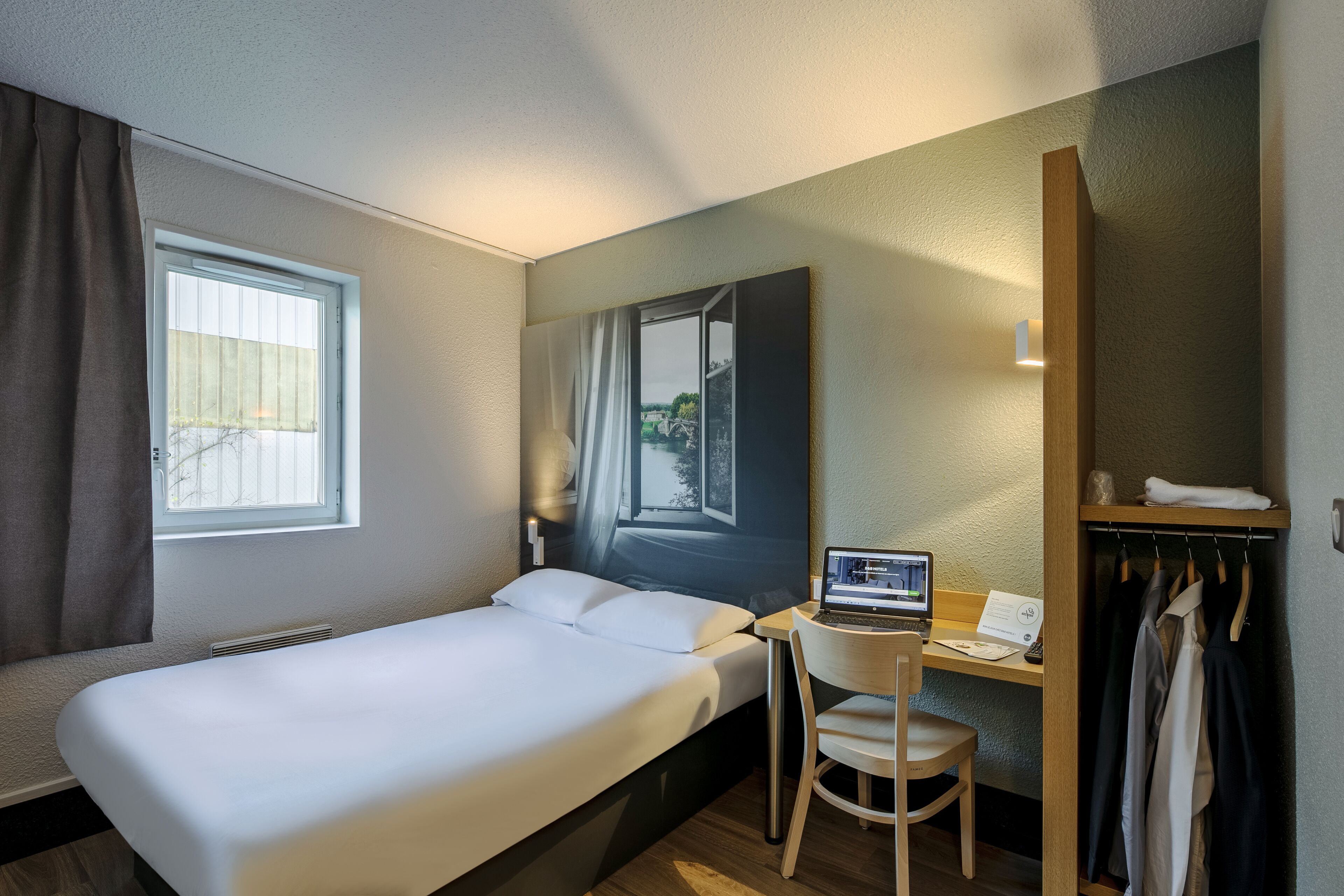A hotel room with a bed, a desk with a laptop, a chair, a wardrobe, and a window with curtains.