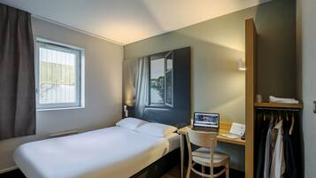 A hotel room with a bed, a desk with a laptop, a chair, a wardrobe, and a window with curtains.