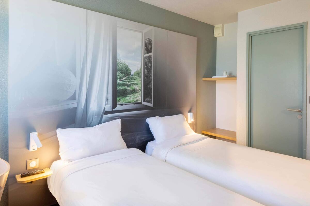 A hotel room with two beds, a window with a view of greenery, a door, and a nightstand with a remote control.