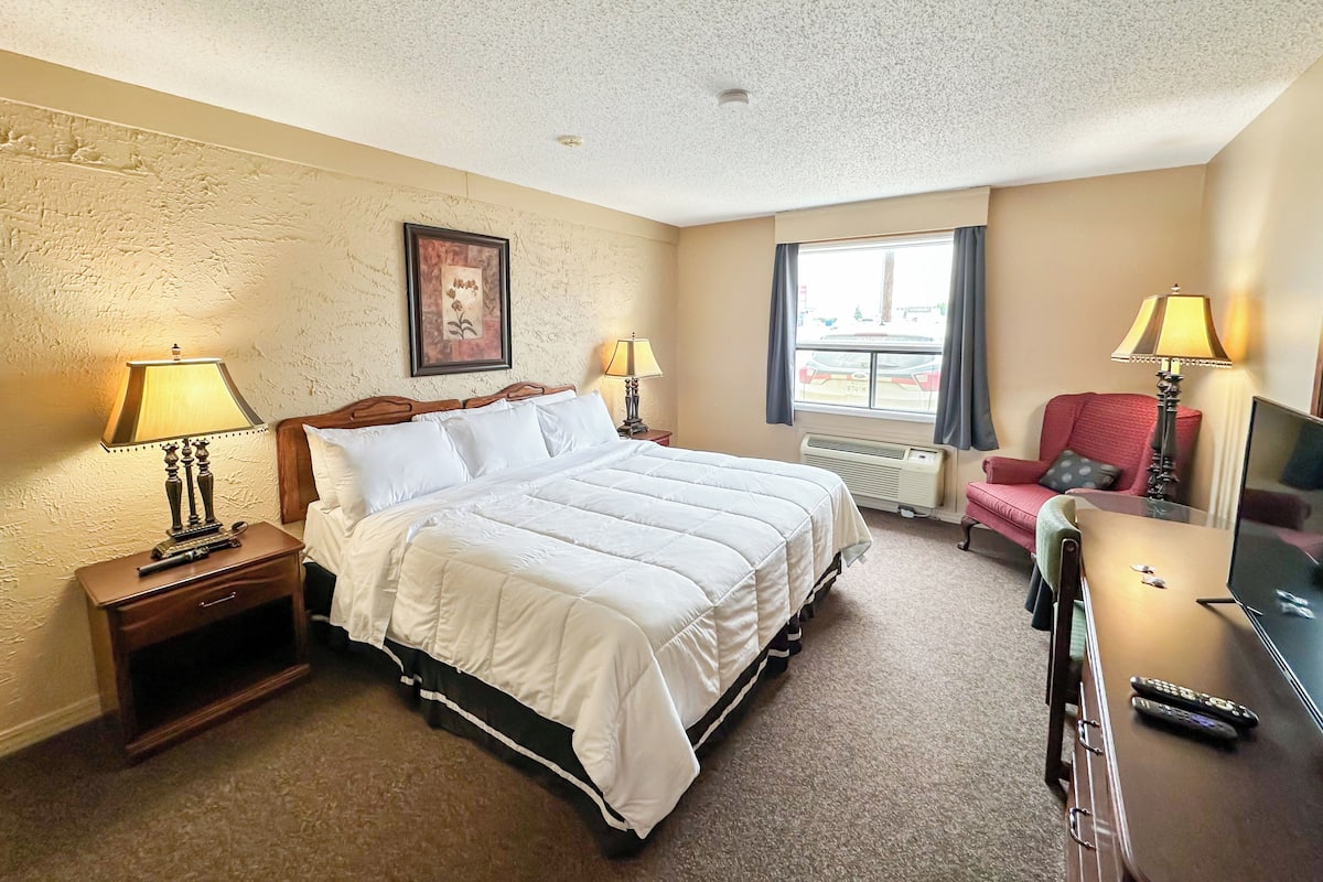 A hotel room with a bed, two bedside tables with lamps, a chair, a TV, and a window with curtains.