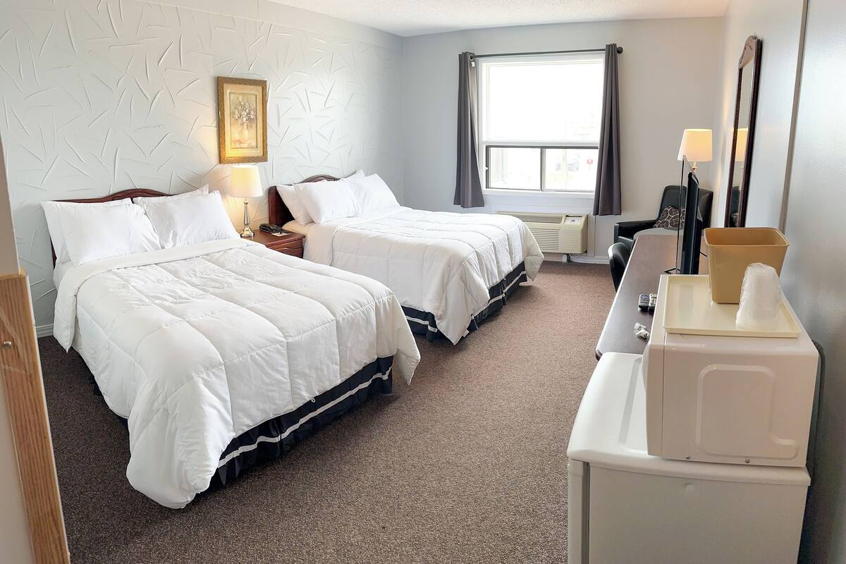 A hotel room with two beds, a desk with a lamp, a window with curtains, and a mirror.