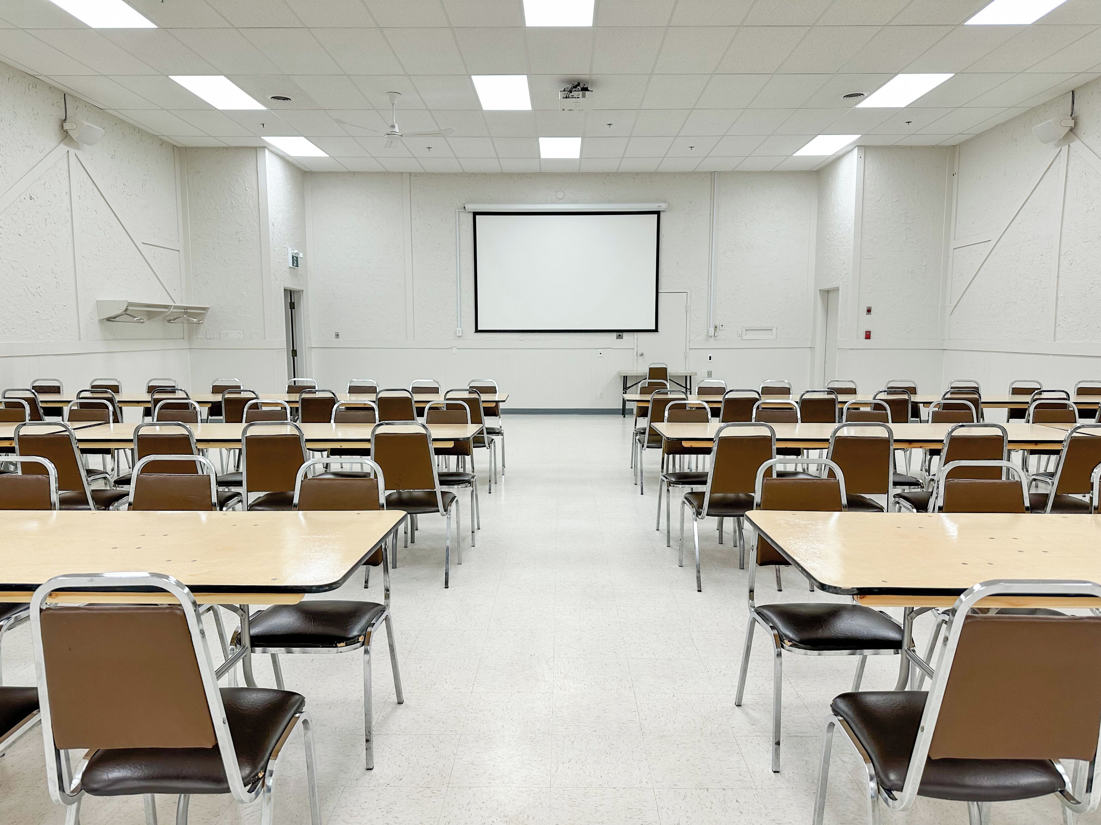 A classroom with rows of desks and chairs, a projector screen, and a ceiling fan.