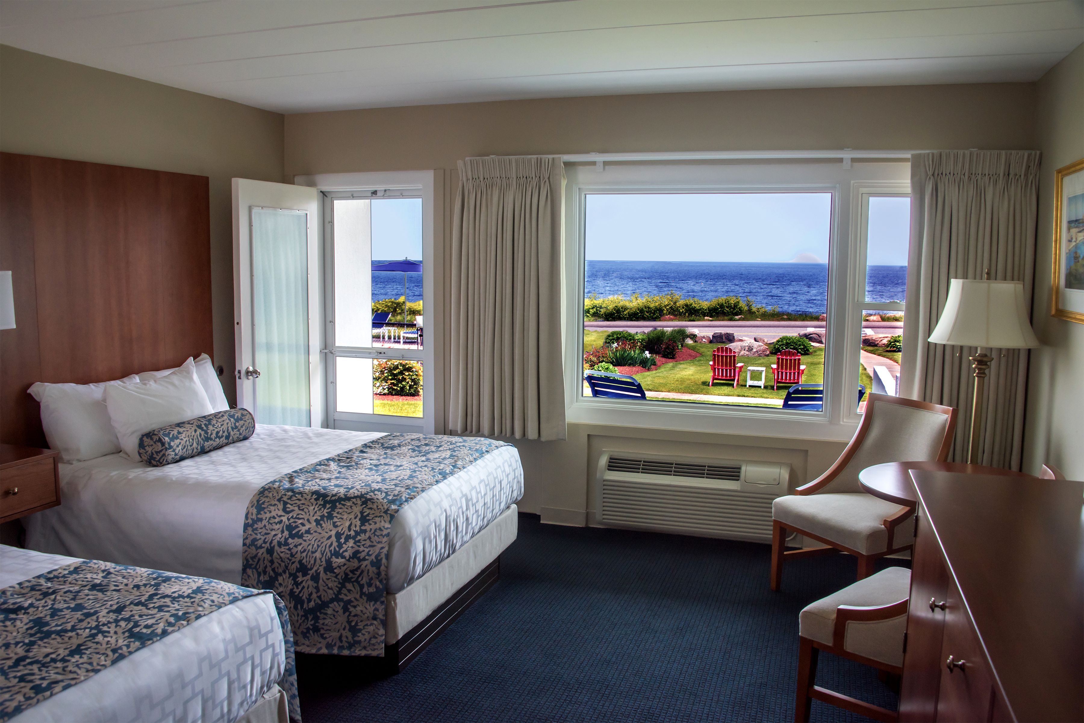 A hotel room with two beds, a desk, and a chair. The room has large windows with a view of the ocean and a distant mountain.
