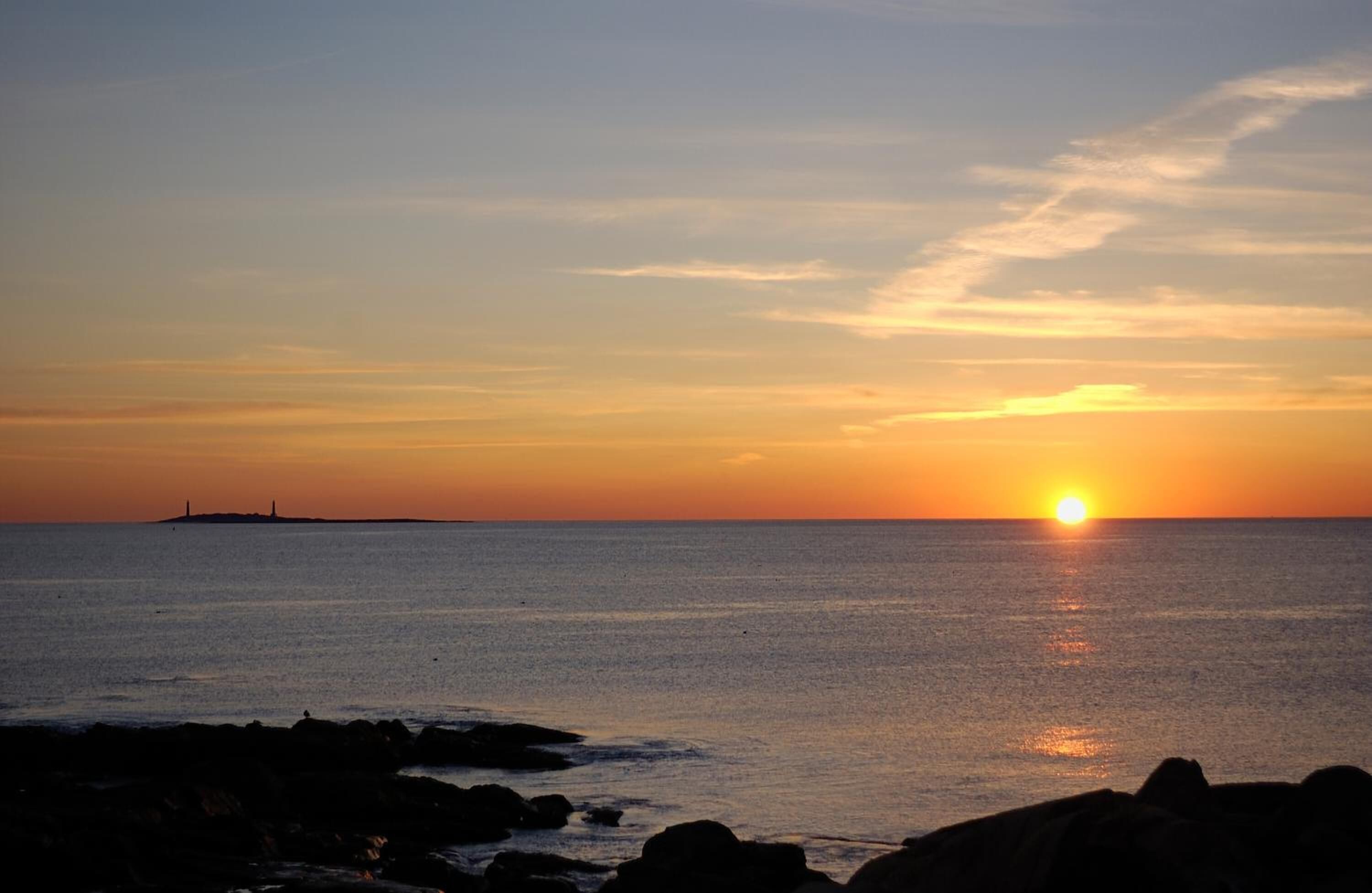 A sunset over the ocean with a distant island and rocky shoreline.