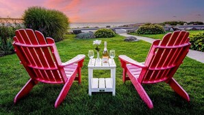 Zwei rote Adirondack-Stühle stehen einem weißen Tisch mit einer Flasche und Gläsern gegenüber, platziert auf einer grünen Rasenfläche mit Blick auf das Meer und einen Sonnenuntergang.