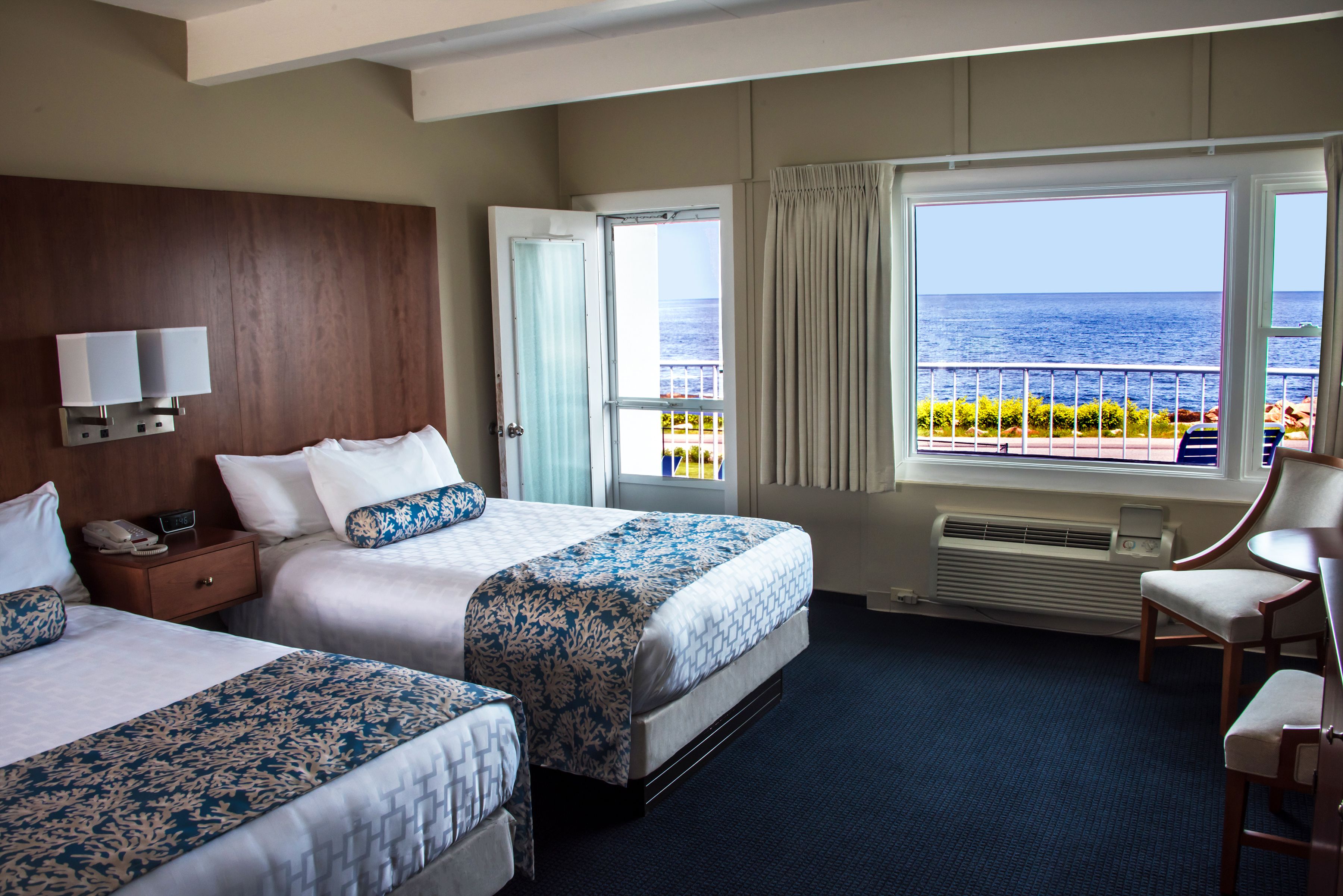 A hotel room with two beds, a nightstand, a chair, a large window with a view of the sea, and a wall-mounted lamp.