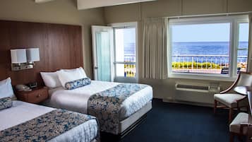 A hotel room with two beds, a nightstand, a chair, a large window with a view of the sea, and a wall-mounted lamp.