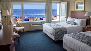 Ein Hotelzimmer mit zwei Betten, einem Schreibtisch und einem Sessel. Ein großes Fenster bietet Blick auf den Ozean, und ein Balkon ist vorhanden.