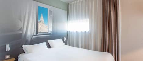 A hotel room with a large bed, a nightstand, a window with curtains, and a view of a building outside.